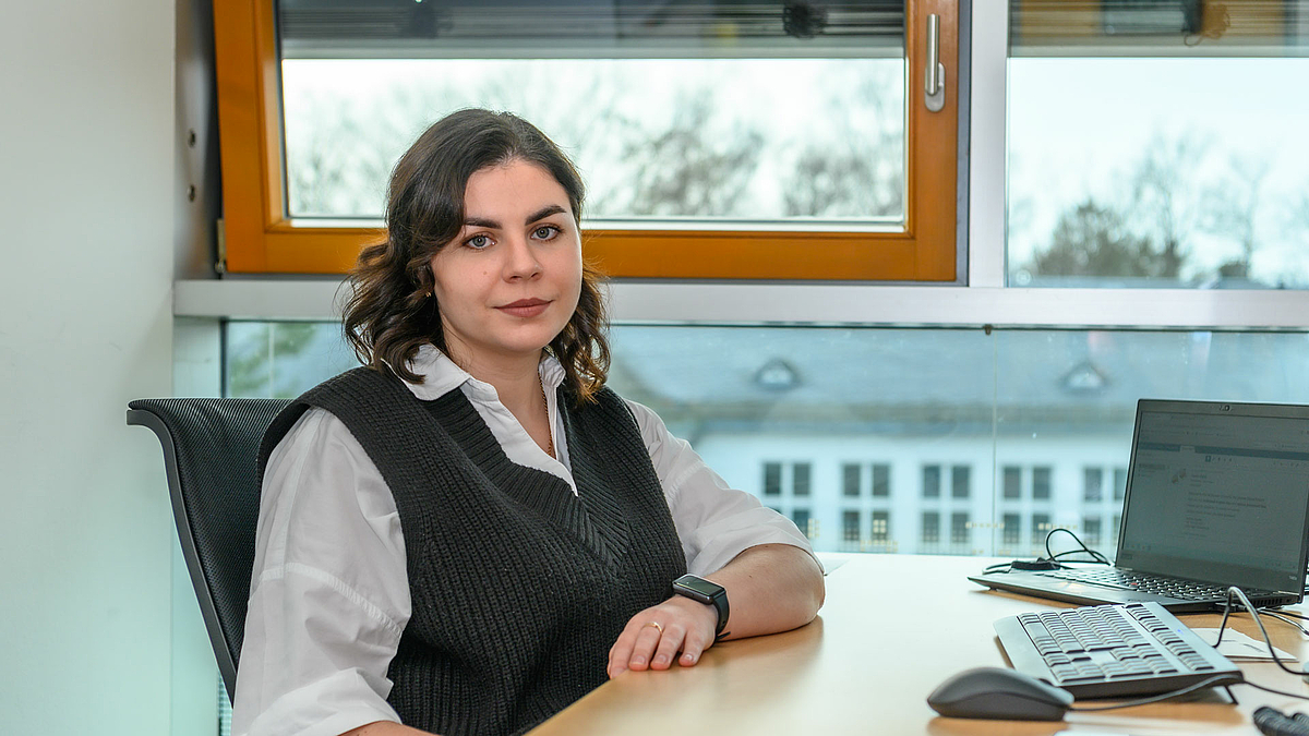 Daryna Karpiuk ist ukrainische Studentin und arbeitet in einem der Projekte der HSMW zur Unterstützung ihrer Landsleute. Daryna Karpiuk sitzt an einem Schreibtisch mit Laptop vor einer Fensterfront. Sie trägt Bluse und Wollweste und blickt freundlich, aber bestimmt in die Kamera.