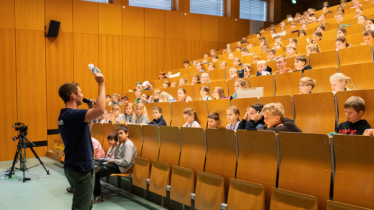 Moderator steht unten vor den Kindern in den ansteigenden Bankreihen im Hörsaal und hält ein Objekt hoch.