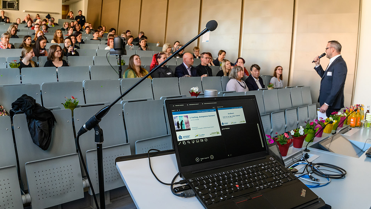 Blick in den Hörsaal mit den Teilnehmenden in ansteigenden Bankreihen. Auf einem Laptop im Bildvordergrund ist die Begrüßungsfolie der Fachtagung zu sehen. Neben dem Laptop stehen blühende Pflanzen. Rechts im Bild ist eine männliche Person mit Mikrofon in der Hand, die zu den Teilnehmenden im Hörsaal spricht.