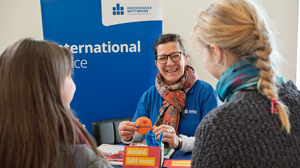 Lachende weibliche Person vor "INternational-Office" Rollup hält ein "I love Erasmus+"-Kärtchen in den Händen und unterhälts ich mit zwei Personen im Bildvordergrudnd