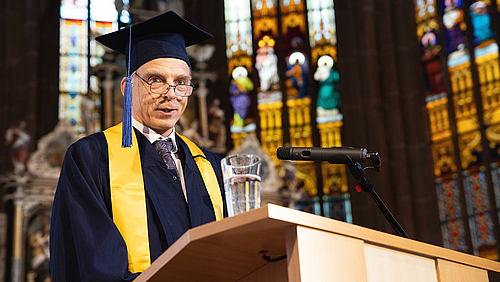 „Ich immatrikuliere Sie in die Gemeinschaft der Hochschule für Angewandte Wissenschaften Mittweida.“ Prof. Dr. Volker Tolkmitt immatrikulierte die Erstsemester des Wintersemesters 2022/2023 feierlich. Volker Tolkmitt spricht am Pult im Altarraum der Mittweidaer Stadtkirche. Er trägt einen dunkelblauen Talar und Hut. Im Hintergrund fällt Sonne durch die farbenfrohen Kirchenfenster.