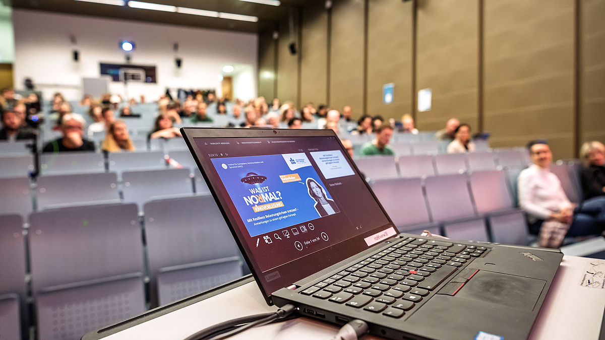 Ein aufgeklapptes Laptop mit der Begrüßungsfolie zur Reihe „Was ist normal?“ auf dem Display. Im Hintergrund sich unscharf die Zuhörer:innen in den ansteigenden Bankreihen des Hörsaals zu sehen.