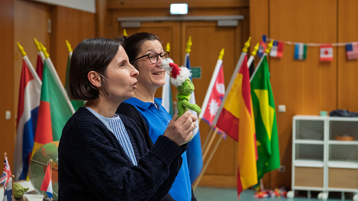 Zwei Dozentinnen stehen nebeneinander, eine hält eine Grinch-Figur, im Hintergrund mehrere Länderflaggen.