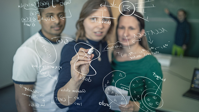 Zwei Frauen und ein Mann stehen vor einer Glasscheibe, auf die sie mathematische Formeln schreiben. Im Hintergrund steht ein Professor an einer klassischen Tafel.