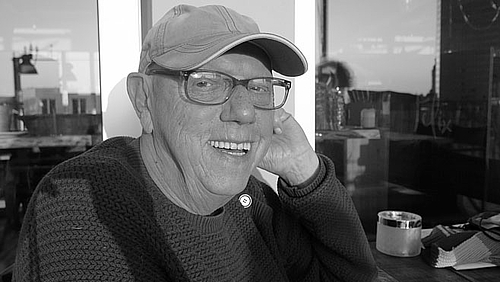 Wolfgang Scherer, 1947 – 2022 Das Schwarzweißfoto zeigt den Verstorbenen. Er trägt Basecap und Strickpullover und lacht fröhlich in die Kamera.