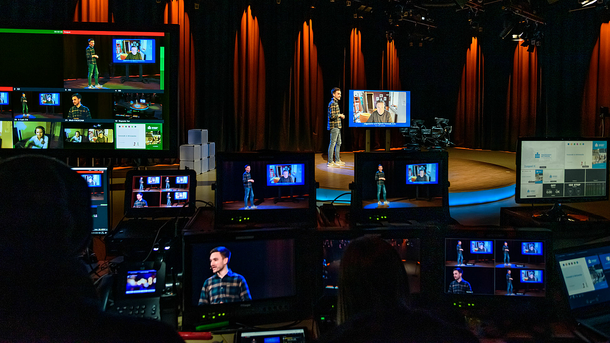 Auf dem Foto sind im Vordergrund zahlreiche Monitore zu sehen, davor sind im Dunkeln und von hinten Köpfe zu erkennen. Die Monitore zeigen zum Teil das Motiv das sich im Studio im Bildhintergrund abspielt: ein Moderator auf einem niedrigen runden Podest und neben ihr stehend ein großer Monitor, über den eine weitere Person zugeshaltet ist.