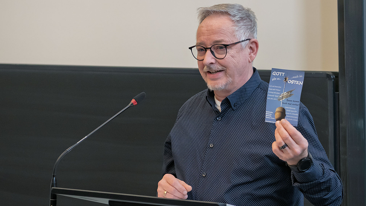 Männliche Person am Rednerpult mit Flyer in der Hand, auf dem unter anderem „Gott gibt es auch im Osten“ zu lesen ist.