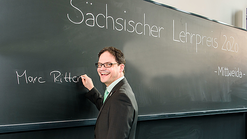 Professor Marc Ritter ganz klassisch. Den Sächsichen Lehrpreis bekam er heute für ein innovatives Konzept, das Präsenz- und Onlinelehre verbindet. Das Fotos zeigt den Preisträger Marc Ritter, der gerade seinen Namen an die Kreidetafel eines Hörsaals gerschrieben hat.