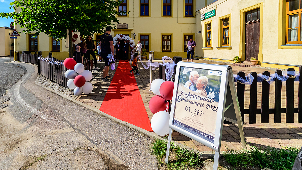 Ein roter Teppich führt vom Vorplatz des Gasthauses zum Eingang im Hintergrund, der mit roten und weißen Ballons gescmückt ist. Im Vordergrund steht ein Aufsteller mit dem Seniorenball-Plakat.