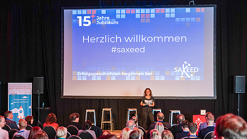 Erfolgsgeschichte schreibt man mit Erfolgsgeschichten. Die Geburtstagsfeier moderiert von Jenny Spanka stellte viele davon vor. Auf einer Bühne steht eine weibliche Person und spricht zu zahlreichen sitzenden Zuschauern, deren Köpfe von hinten im Bildvordergrund zu sehen sind. Auf der Leinwand hinter der Bühne ist eine Grafik projiziert, auf der auf blauem Grund „15+1 Jahre Jubiläum“, „Herzlich willkommen #saxeed“, „Erfolgsgeschichten beginnen bei SAXEED“ zu lesen ist.