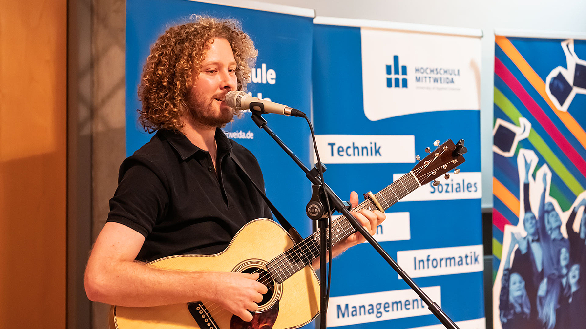 Männliche Person in Poloshirt mit Gitarre singt stehend am Mikrofon vor den drei blauen Rollups der Hochschule.