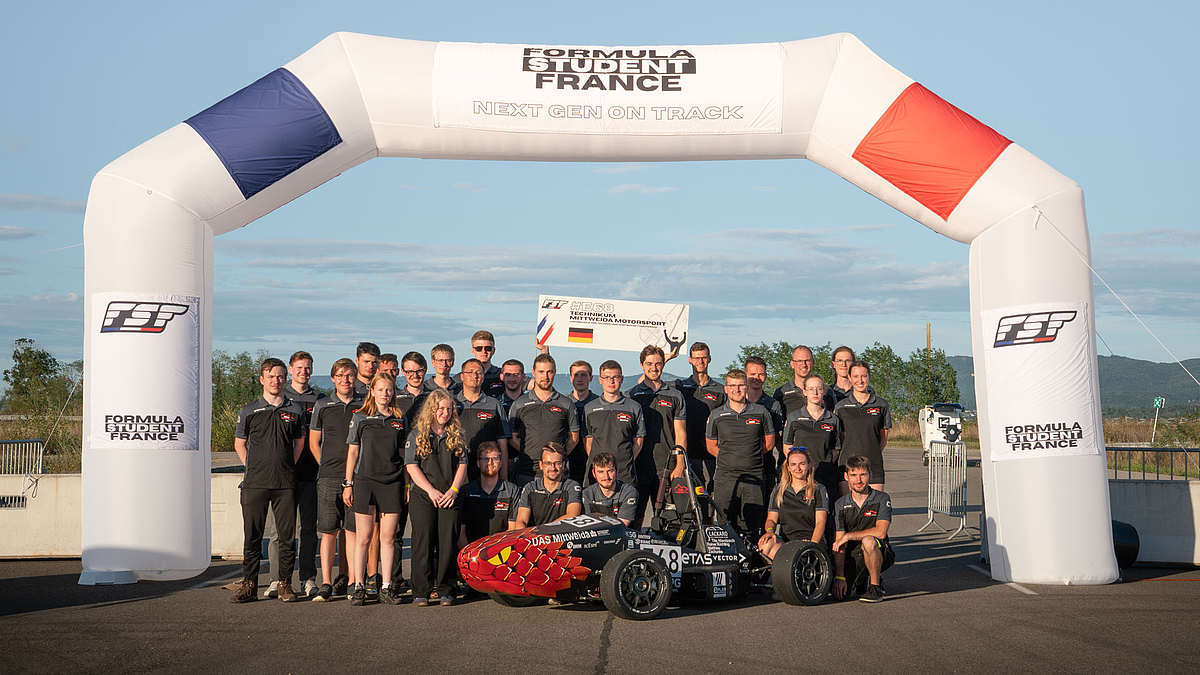 30 junge Menschen in TMM-Polos und ein Rennauto unter einem aufblasbaren Torbogen mit "Formula Student France"-Aufdruck.er einem htgt
