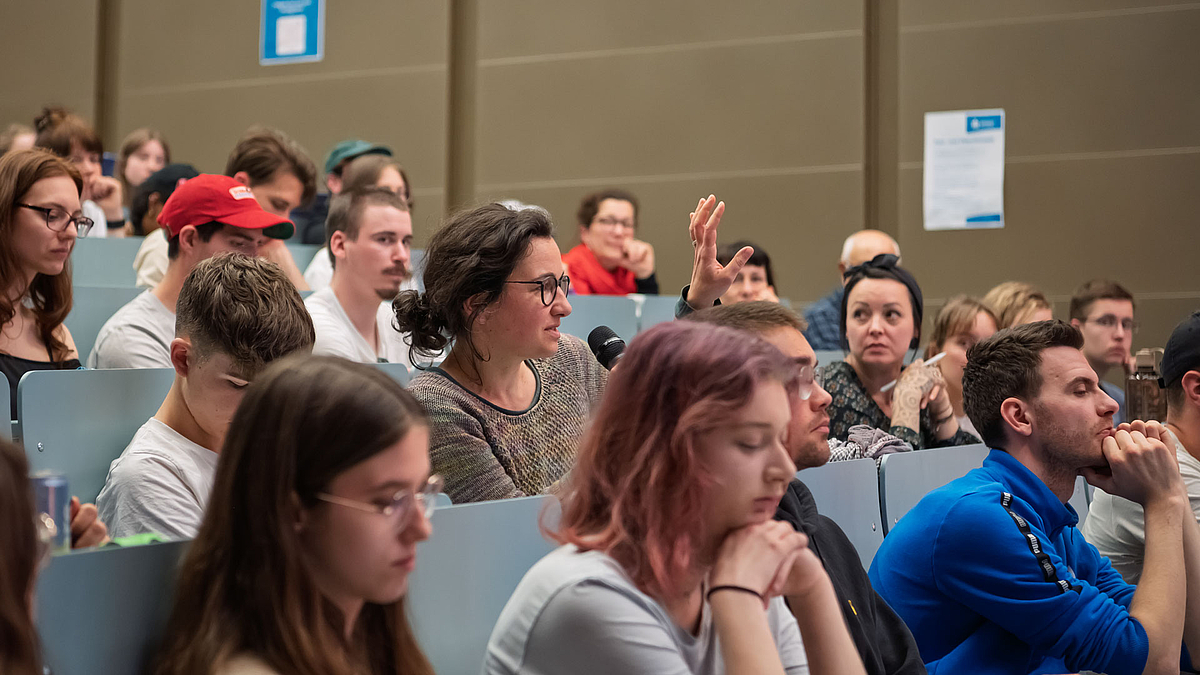Personen im Hörsaal. In der Bildmitte eine weibliche Person, die in ein Handmikrofon spricht und die andere Hand hochhält.