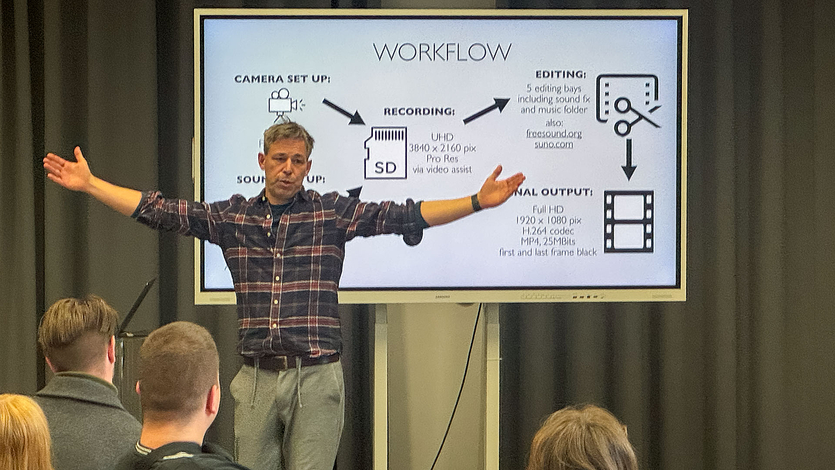 Nähere Einsstellung auf den Dozenten. Auf dem Monitor hinter ihm ist ein mit "Workflow" überschriebenes Schauibild zu sehen.