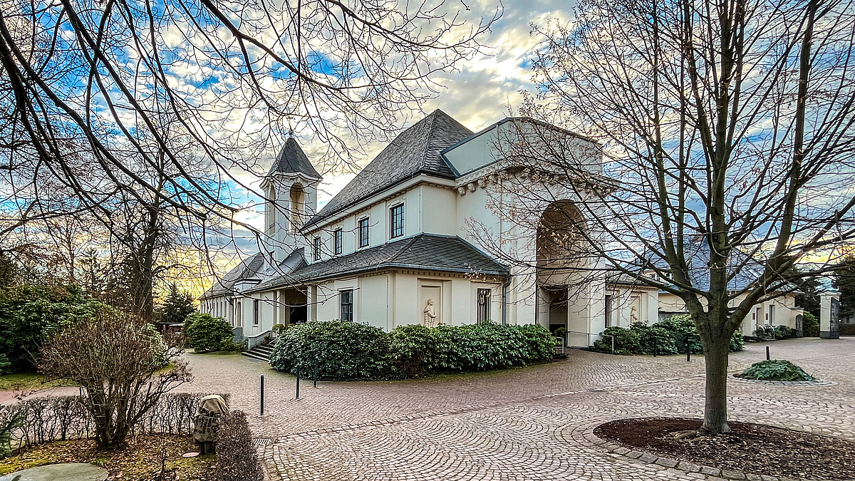 Die im Jahr 1919 fertigestellte Friedhofskapelle des Neuen Friedhofs von Mittweida. Blick auf ein vor einem in der Abendsonne strahlenden, hellen kirchenartiges Gebäude mit Turm und großem Engangsportal in einer parkartigen Umgebung mit Bäumen und Sträuchern.
