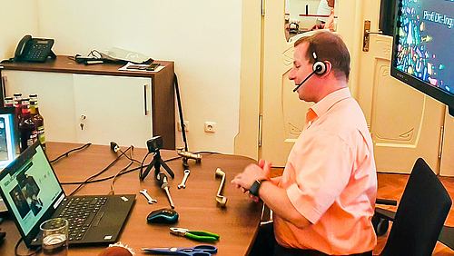 Professor Uwe Mahn hat zur Kinderuni viele Alltagsgegenstände mitgebracht, an denen sich das Thema "Alles Hebel" gut demonstrieren lässt. Das Foto zeigt den Dozenten Porfessor Uwe Mahn an einem Tisch. Eine Webcam ist auf ihn gerichtet. Auf dem Tisch liegen unter anderem eine Schere, eine Türklinke und mehere Schraubenschlüssel, an denen sich das Thema "Alles Hebel" demonstrieren lässt.