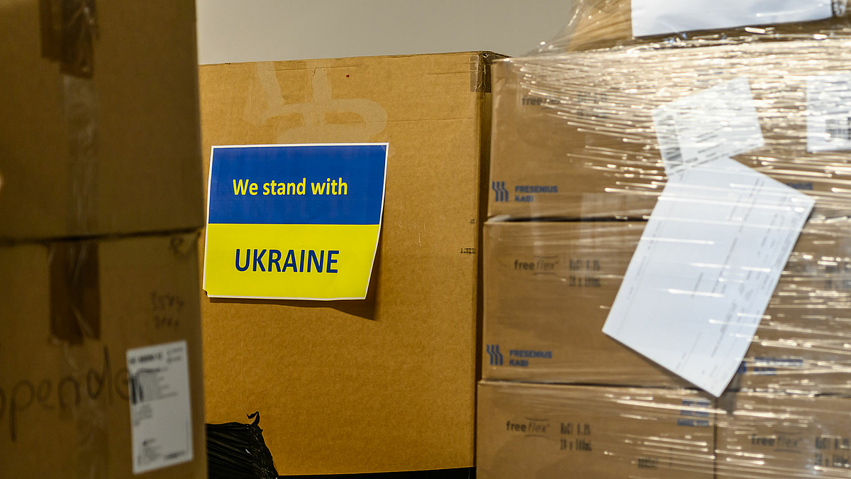 Das Foto zeigt kleines Plakat in den Farben der ukrainischen Nationalflagge mit der Aufschrift „We stand with“ (oben gelb auf blau) „Ukraine“ (unten blau auf gelb).