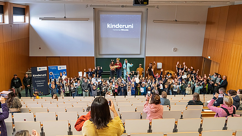 Diplomiert und fotografiert: Abschluss des Kinderuni-Jahres an der Hochschule Mittweida mit Urkunden und stolzen Eltern. Hörsaal mit ansteigenden Bankreihen. Unten stehen rund einhundert fröhliche Kinder mit Urkunden. Hinter ihnen und Teil auf dem Pult stehe die Mitglieder des Kinderuni-Teams in ihren Tier-Kostümen. In den Bankreihen stehen Eltern, die mit ihren Smartphones Fotos machen.