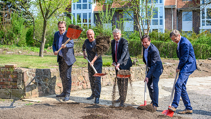 Das Foto zeigt fünf männliche Personen mit Schaufeln, mit denen sie Erde aufgenommen haben, die in den Bildvordergrund geworfen wird.