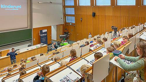 Kinderuni-Organisator Robin Biebl (links) begrüßt viele Kinder im großen Hörsaal der Hochschule Mittweida. Nicht im Bild und doch im Bilde: (Groß-)Eltern schauen vom Nachbarhörsaal aus zu.