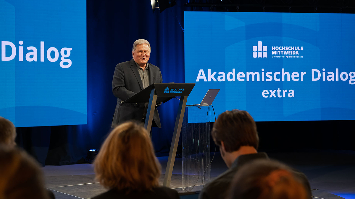 Männlicher Referent am Rednerpult lächelt. Hinter ihm auf einem LED-Screen ist in weißer Schrift vor blauem Hintergrund "Akademischer Dialog extra" unter dem Logo der Hochschule zu lesen.