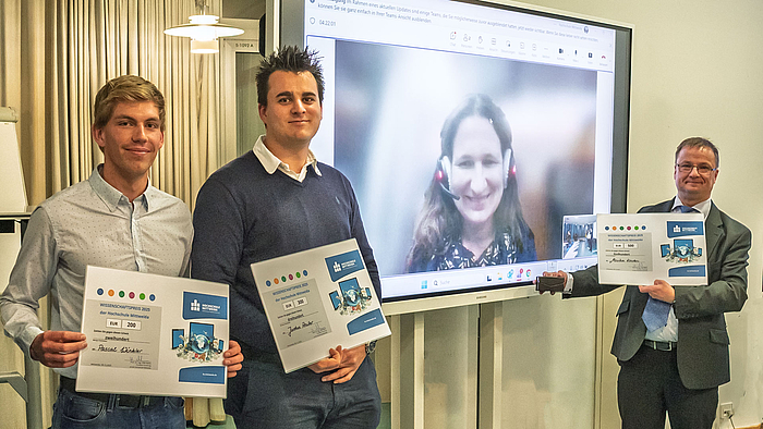 Prorektor Forschung Professor Uwe Mahn (r.) mit den Preisträger:innen des Wissenschaftspreises der HSMW 2025: Pascal Winkler, hier vertreten von Jim Köcher, Justus Reuter und live zugeschaltet Dr. Marika Kaden. Drei männliche Personen mit Urkunden in der Hand. Zwischen ihnen auf einem großen Bildschirm eine weibliche Person.