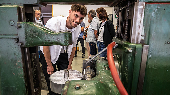 Max Brunner hat den Durchblick – im doppelten Sinn des Wortes. Fast sechs Monate hat der Maschinenbaustudent neben seinem Studium daran gearbeitet, die über siebzig Jahre alte Wälzfräsmaschine flott zu bekommen. Eine junge männliche Person blickt lächelnd durch eine alte Maschine mit Zahnrädern, Wellen und Ketten hindurch. Im Bildhintergrund stehen weitere Personen.