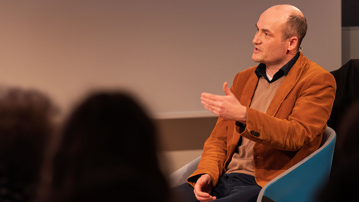 Podiumsteilnehmer sitzend und freundlich gestikulierend. Im Vordergrund sich unscharf Köpfe von Zuschauenden zu sehen.