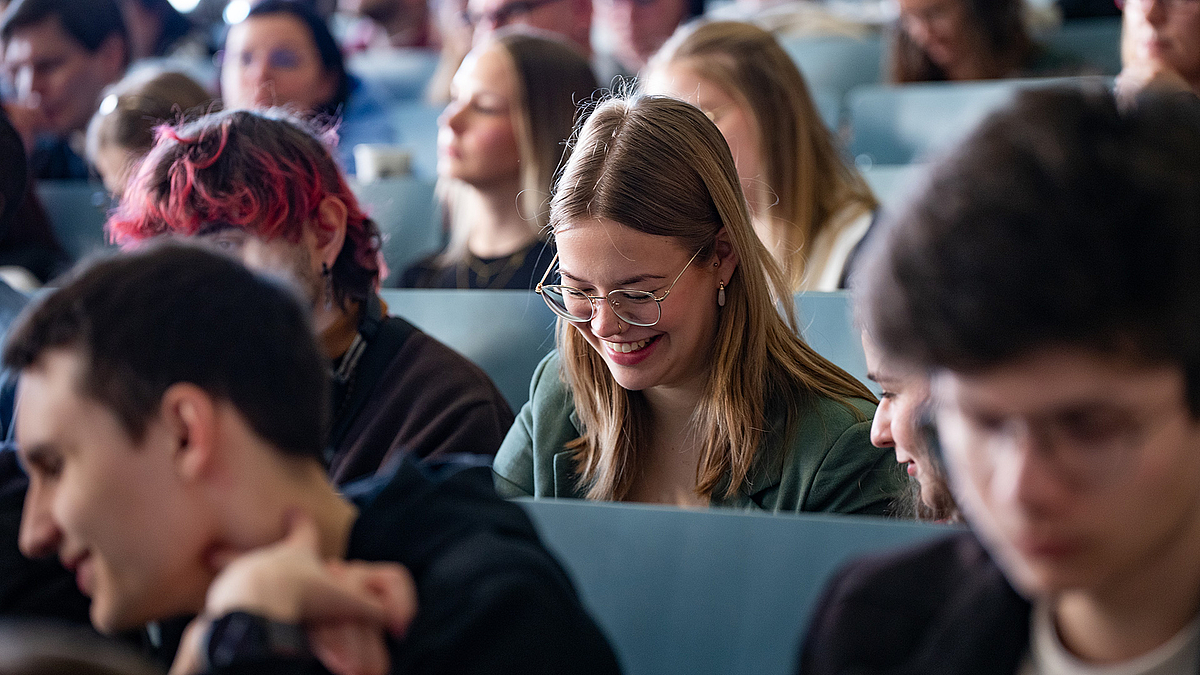 Insgesamt liegt der Anteil der neuen Studentinnen im Sommersemester bei 74 Prozent, der Gesamtanteil weiblicher Studierender unter allen aktuell 5244 Immatrikulierten der HSMW beträgt rund 37 Prozent. Eine Studentin sitzt im Hörsaal. Sie schaut nach unten und grinst breit mit offenem Mund. Ganz am rechten Bildrand sieht man nur versteckt, dass eine Kommilitonin zu ihr gewandt ist und offenbar mit ihr redet.