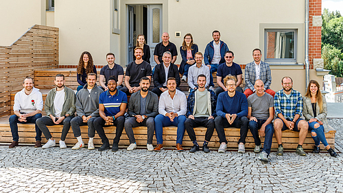 Pause beim Pitch für Teilnehmende und die Jury: Studierende des Blockchain-Masterstudiengangs der Hochschule Mittweida präsentieren zum Semesterabschluss ihre Geschäftsideen. 22 Personen unterschiedlichen Alters und Geschlechts sitzen bei schönem Wetter und guter Stimmung in drei aufsteigenden Reihen im Freien vor einer Hauswand.