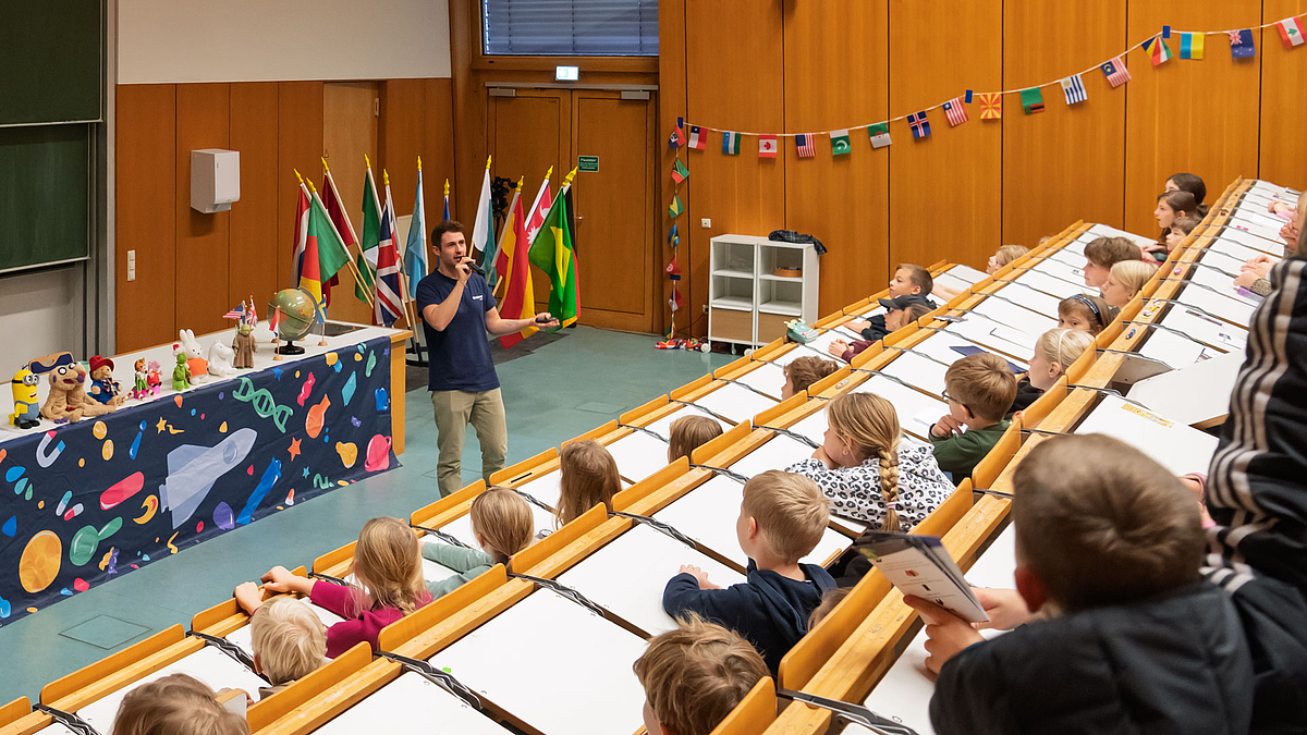 Moderator steht vor Publikum in Hörsaal mit internationalen Flaggen und Vernetzung auf Leinwand.