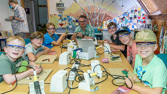 Das Foto zeigt sechs lachende Kinder Eddie, Alexander, Benjamin, Aaron, Dominic und David mit Falko Jahn von Laserinstitut Hochschule Mittweida (Mitte) an einem großen Tisch mit Lötstationen. 