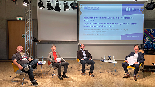 Richard Börner (r.) moderierte beim Netzwerktreffen der Mathematiker und Physiker die Podiumsdiskussion. Vier Personen sitzen auf Stühlen in einem großen Raum und werden von Decken-Scheinwerfern beleuchtet. Im Hintergrund ist eine Powerpoint-Folie auf eine Leinwand projiziert, die den Titel „Digitale Lehre nach(?) Corona – lesson learned oder zurück auf Start?“ trägt.