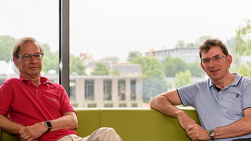 Laserkompetenz kombiniert: Prof. Dr. rer. nat. Steffen Weißmantel (links) und Prof. Dr. rer. nat. habil. Alexander Horn leiten die DFG-geförderte Grundlagenforschung im Projekt MULTIPULS. Das Foto zeigt zwei männliche Personen mittleren Alters auf einem Sofa vor einer großen Fensterfläche: Prof. Steffen Weißmantel und Prof. Alexander Horn. Beider Blick ist dem Betrachtenden zugewandt.