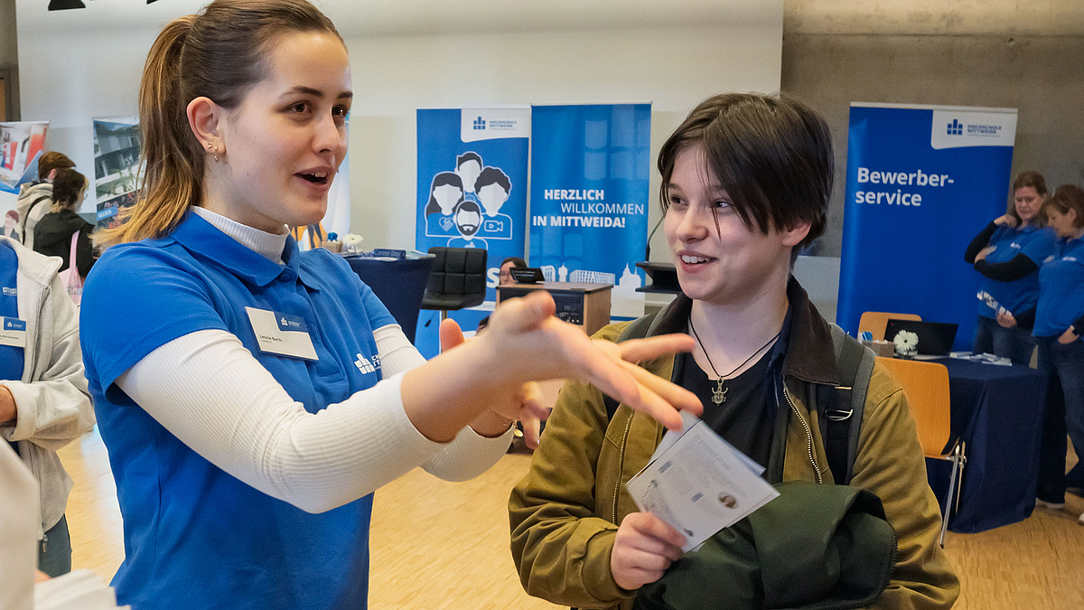 Eine junge weibliche Person im blauen HSMW-Polo mit den Händen gestikulierend und im Gesprräch mit einer weiteren jungen weiblichen Pesron, die Flyer in der Hand hat. Im Hintergrund ist ein Rollup mit der Aufschrift "Willkommen in Mittweida" zu sehen.