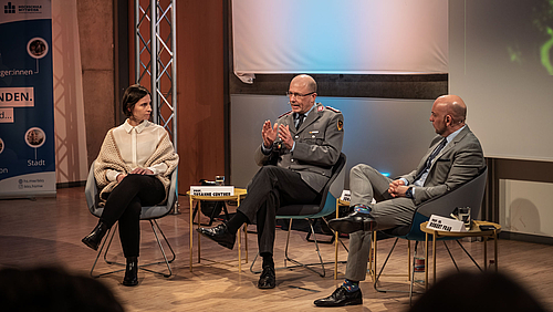 Eine weibliche und zwei männliche Personen in Sesseln im Halbkreis. Die mittlere Person trägt eine Bundeswehruniform. 