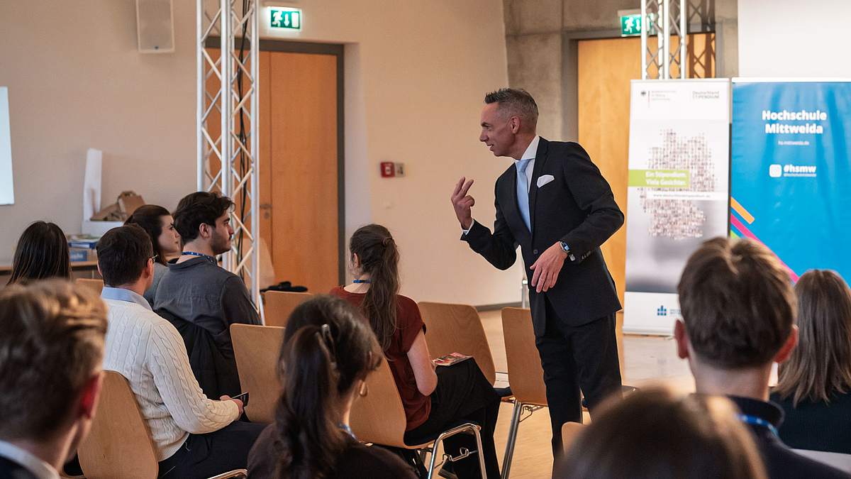 Männliche Person im Anzug agiert vor jungen Leuten an Tischen.