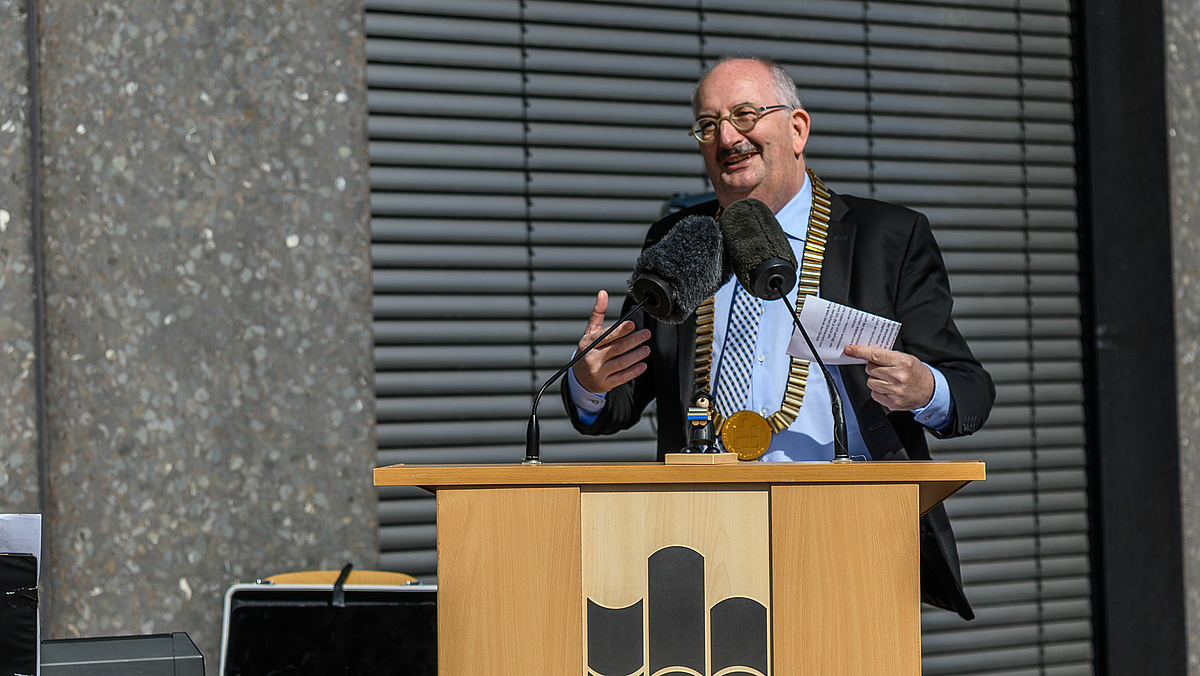 Das Foto zeigt einen Mann mit goldener Amtskette am Rednerpult. An der Vorderseite des Pults ins angeschnitten das Logo der Hochschule zu erkennen.