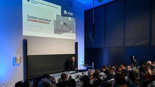 Moderator PD Dr. Gunter Süß, Studiendirektor des Instituts für Kompetenz, Kommunikation & Sprachen (IKKS) der Hochschule Mittweida, sieht sich zum Auftakt der Ringvorlesung im Sommersemester 2022 vor einem voll besetzen Hörsaal. Seitlicher Blick von hinten in die ersten Reihen und auf Pult, Tafel und Beamerprojektion im Hörsaal. Projiziert ist die Begrüßungsfolie der Veranstaltung mit dem Text „Zeiten wenden! Aussitzen – Analysieren – Anpacken, daneben das Foto einer jungen Frau, die einen Rückwärtssalto vor einer grauen Betonwand macht.
