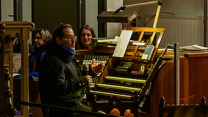 Blick auf den Spieltisch der Orgel mit dme Organisten an den drei Manualen. Er hält ien Mikrofon in der Hand und wendet sich dem Publikum zu.