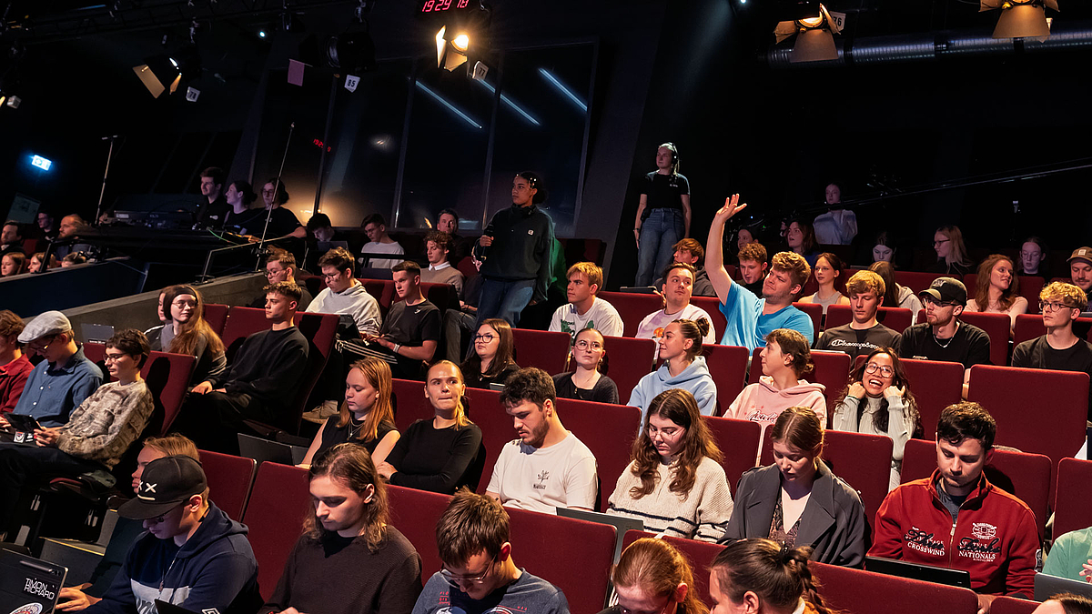 Blick in die ansteigenden Zuschauerränge mit vielen jungen Menschen. Manche schreiben auf Laptops mit. Eine männliche Person meldet sich.