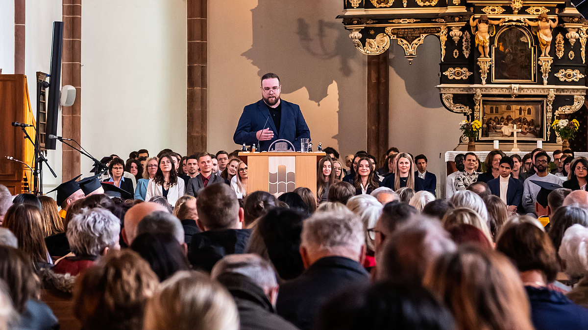 Blick über die Köpfe von Besuchenenden in den Bänken des Kirchensschiffs hinweg auf ein mänliche Person am Rednerpult im Bildhintergrund.
