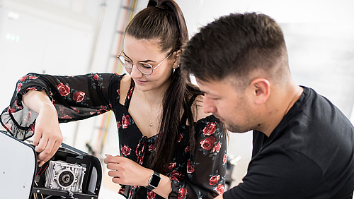 Nie allein: An der Hochschule Mittweida lernen angehende Ingenieur:innen von Dozent:innen mit Industrie-Knowhow. Eine Studentin arbeitet am Inneren eines technischen Geräts, der junge Dozent steht neben ihr zur Unterstützung bereit.
