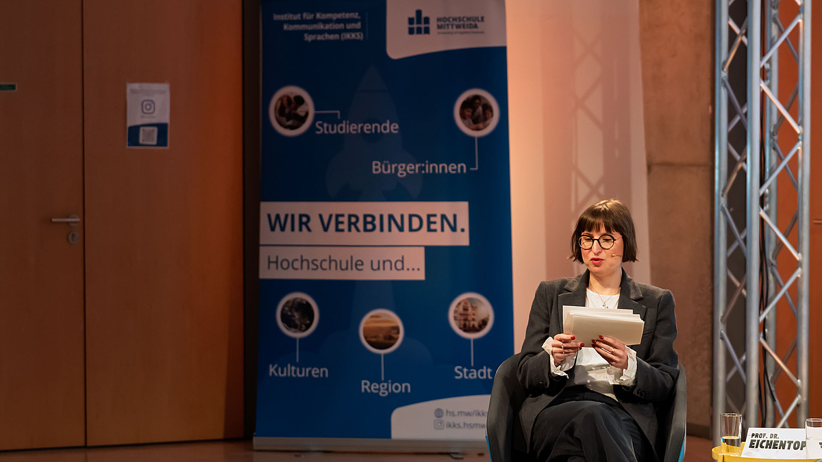 Weibliche Person sitzend mit Moderationskarten. Im Hintergrund ein blaues Rollup unter anderem mit dem Text "Wir verbinden Hochschule und ... ".