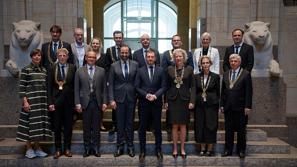 Gruppenbild als Erinnerung für die kommenden acht Jahre: die sächsischen Rektor:innen mit dem aktuellen Ministerpräsidenten und dem Wirtschaftsminister. 14 Männer und Frauen stehen gemeinsam mit Wissenschaftsminister und Ministerpräsident auf einer Steintreppe. Im Hintergrund stehen zwei Löwenplastiken aus hellem Stein.
