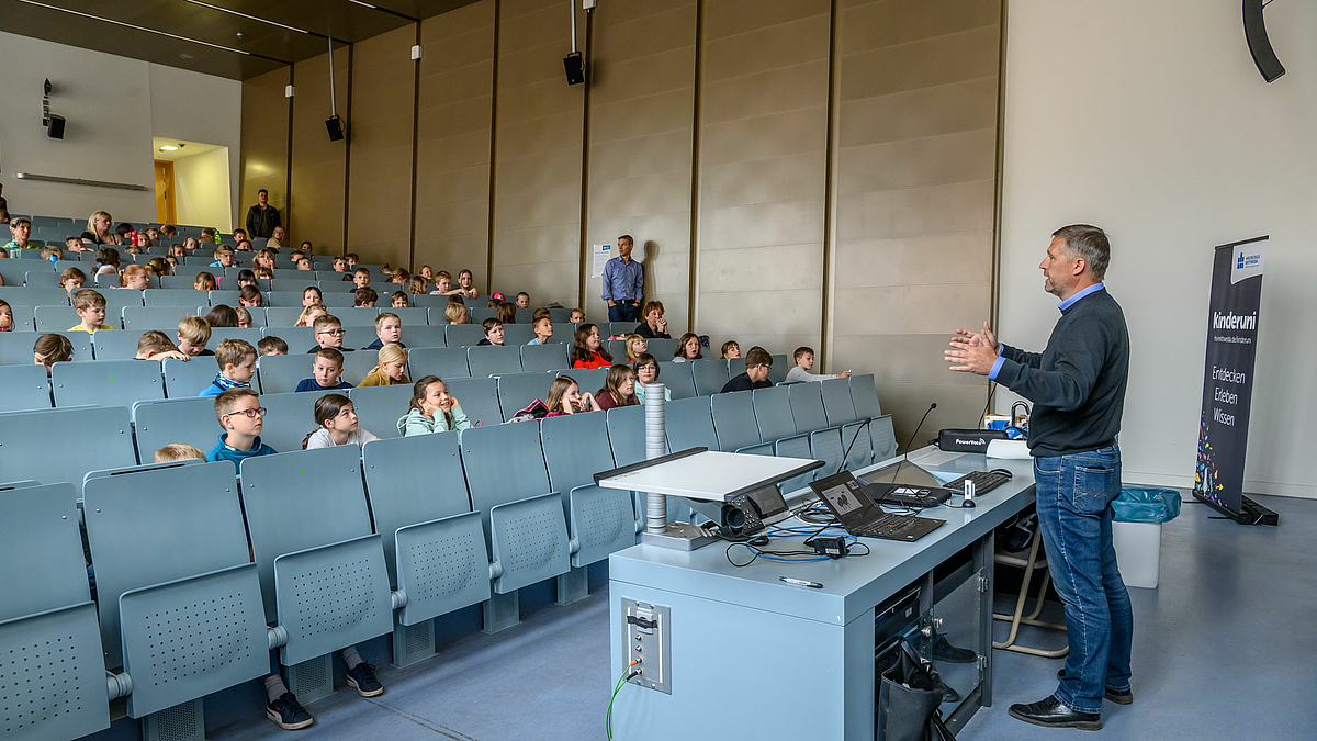 Blick von unten in die aufsteigenden Sitzreihen des Hörsaals in die Gesichter der Kinder. Vorne steht ein Dozent, der gestikulierend zu den Kindern spricht.