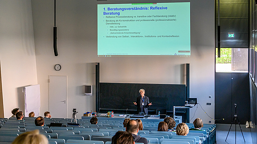 Das Foto zeigt einen Hörsaal mit ansteigenden Sitzreihen von oben fotografiert. Zuhörende sich von hinten zu sehen. Am Pult im Bildhintergrund steht eine männliche Person. Auf die Leinwand oberhalb der Person ist eine Folie projiziert mit der Überschrift "1. Beratungsverständnis: Reflexive Beratunng"