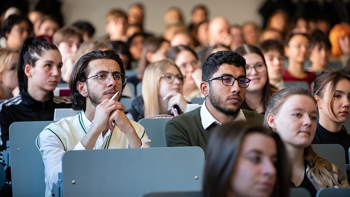 154 Studierende starteten im Sommersemester an der Hochschule Mittweida in einen neuen Lebensabschnitt. 272 bereiten sich als Studienkollegiat:innen auf ihr Studium vor. Zwei junge Männer sitzen auf einer Hörsaalbank und lauschen. Um sie herum sitzen weitere Studierende.