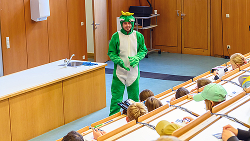 Ein freundlicher Mann im grünen Ganzkörper-Froschkostüm steht vor ienem Pult im Hörsaal. Im Bildvordergrund sitzen Kinder in den ansteigenden Bankreihen.