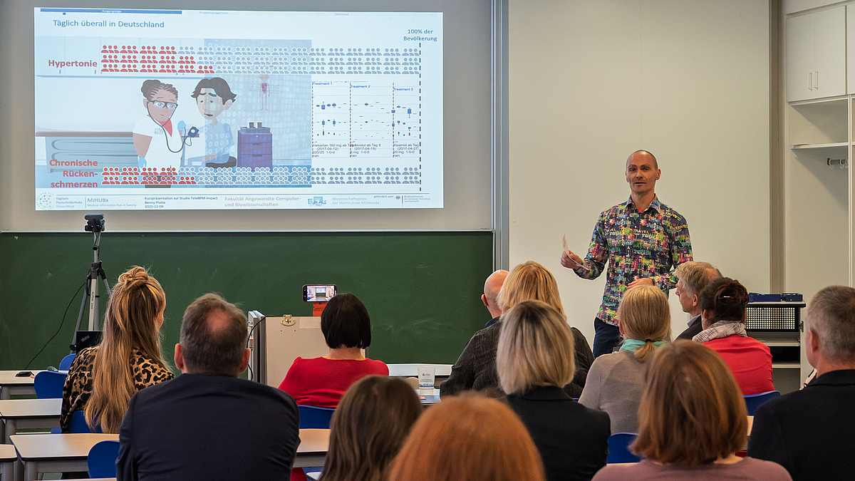 Männliche Person vor projizierter Powerpointpräsentation. Im Vordergrund Köpfe des Publikums von hinten.